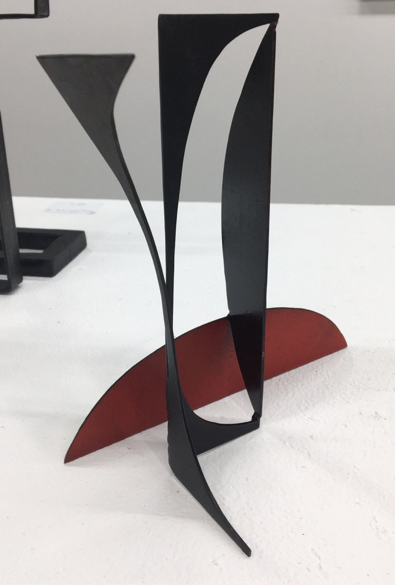 A small sculpture taller than it is wide, made from steel featuring a semi-circular base in a rust red colour with elongated cut out black shapes which rise from it.