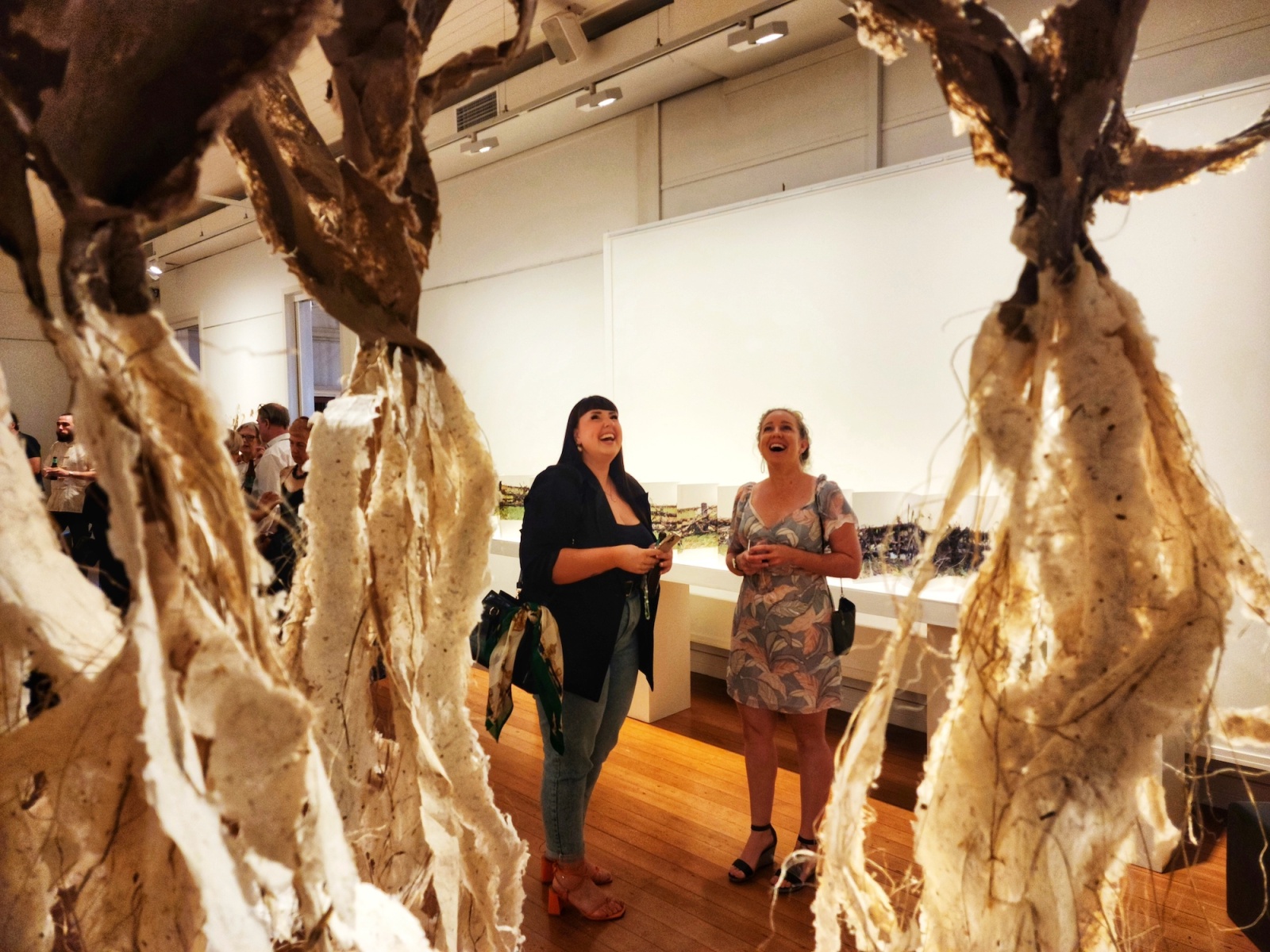 Two women stand in a gallery, smiling and their heads turned to the ceiling; on either side they are flanked by the tendrils of a textural sculpture