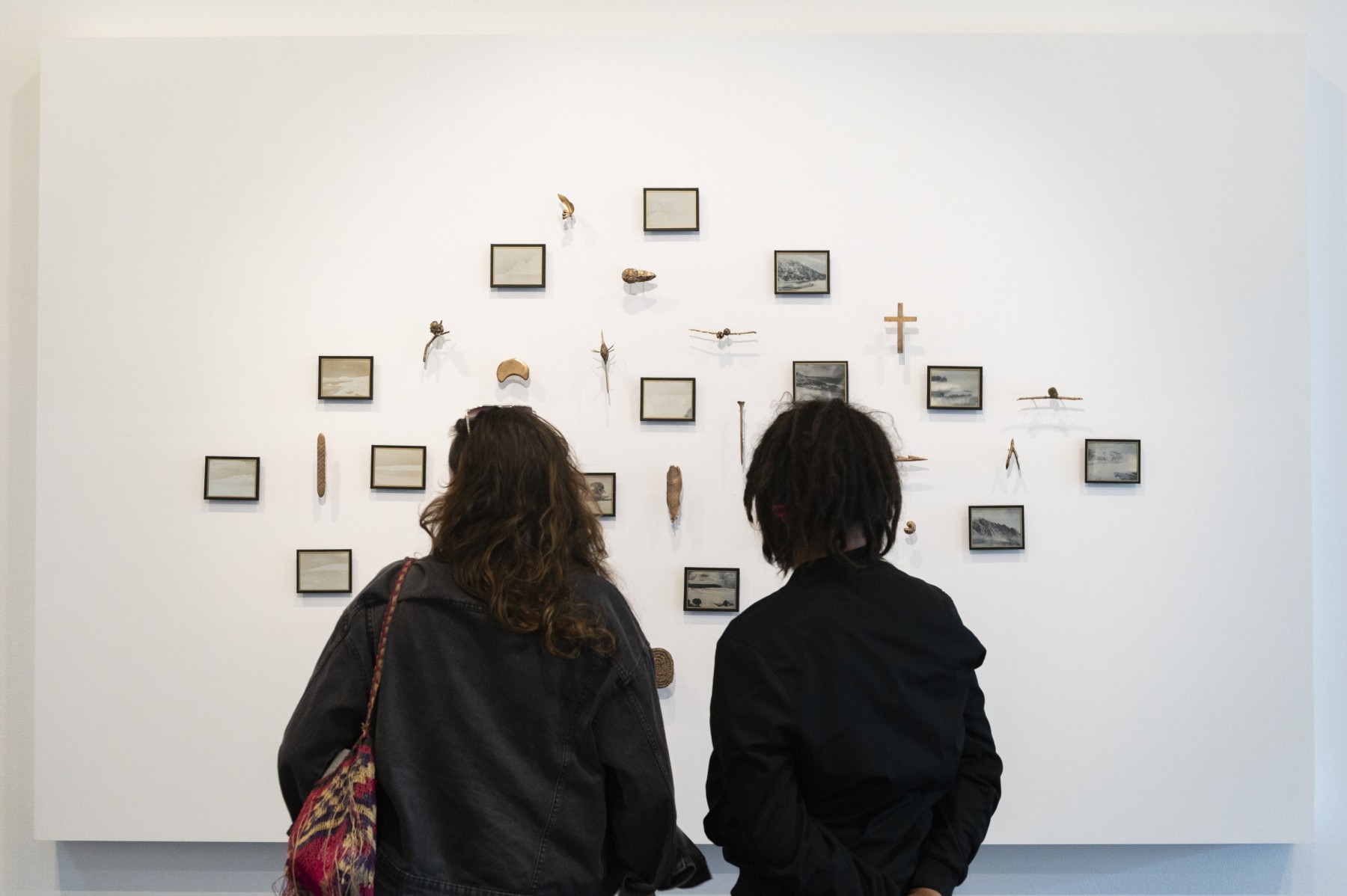 Two figures stand in front of a grid of objects attached to a while wall, they include framed photographs, a Christian cross and Indigenous tools