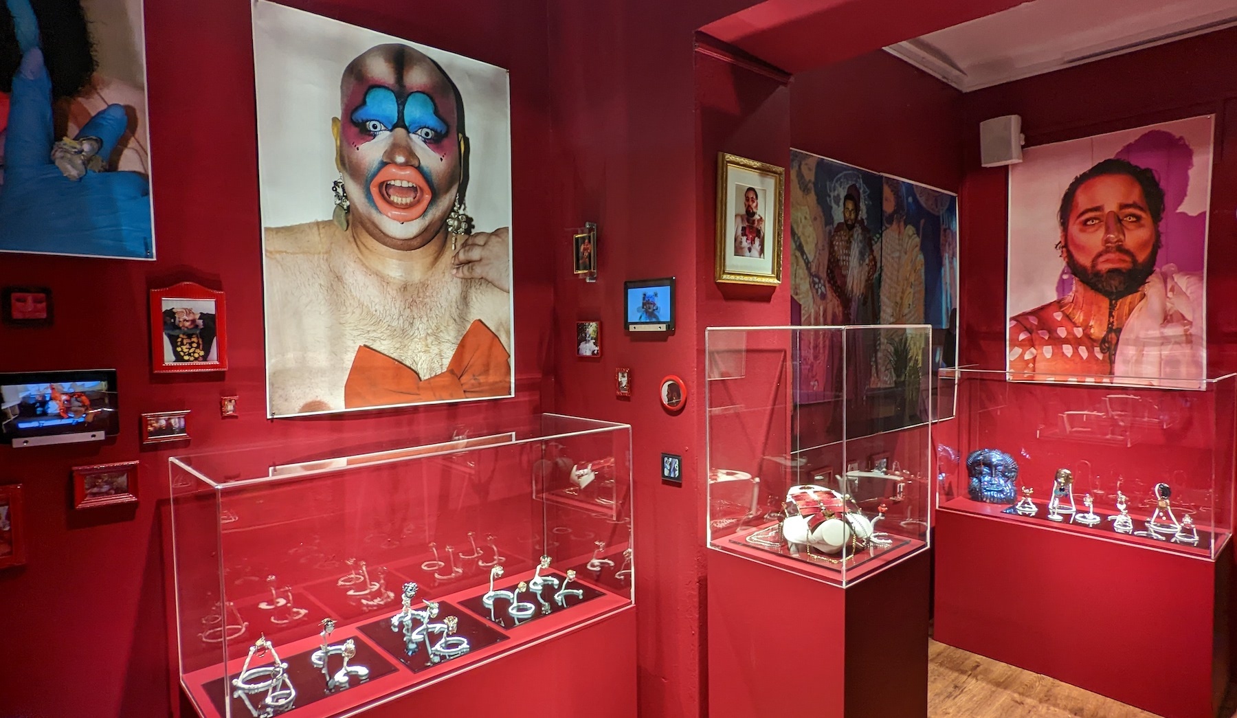 A room with deep red walls is filled with small and large photographs, screens and perspex vitrines holding extravagant jewellery