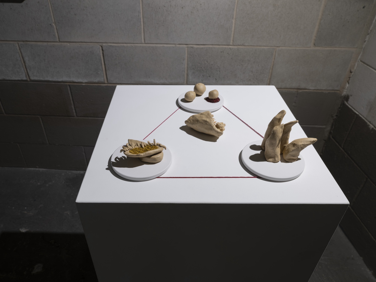 Small clay sculptures sit on top of a white plinth, with red thread connecting them. The plinth sits in the corner of a dimly lit, Besser block room.