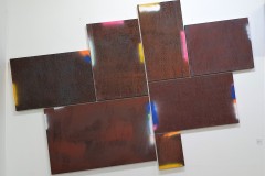 Seven rectangular canvases of different sizes are arranged in a tetris-like grid and angled to the left on a white gallery wall, each canvas has the same richly textured appearance of rust while the edges are variously marked with bright slivers of pink, yellow, white, orange and blue
