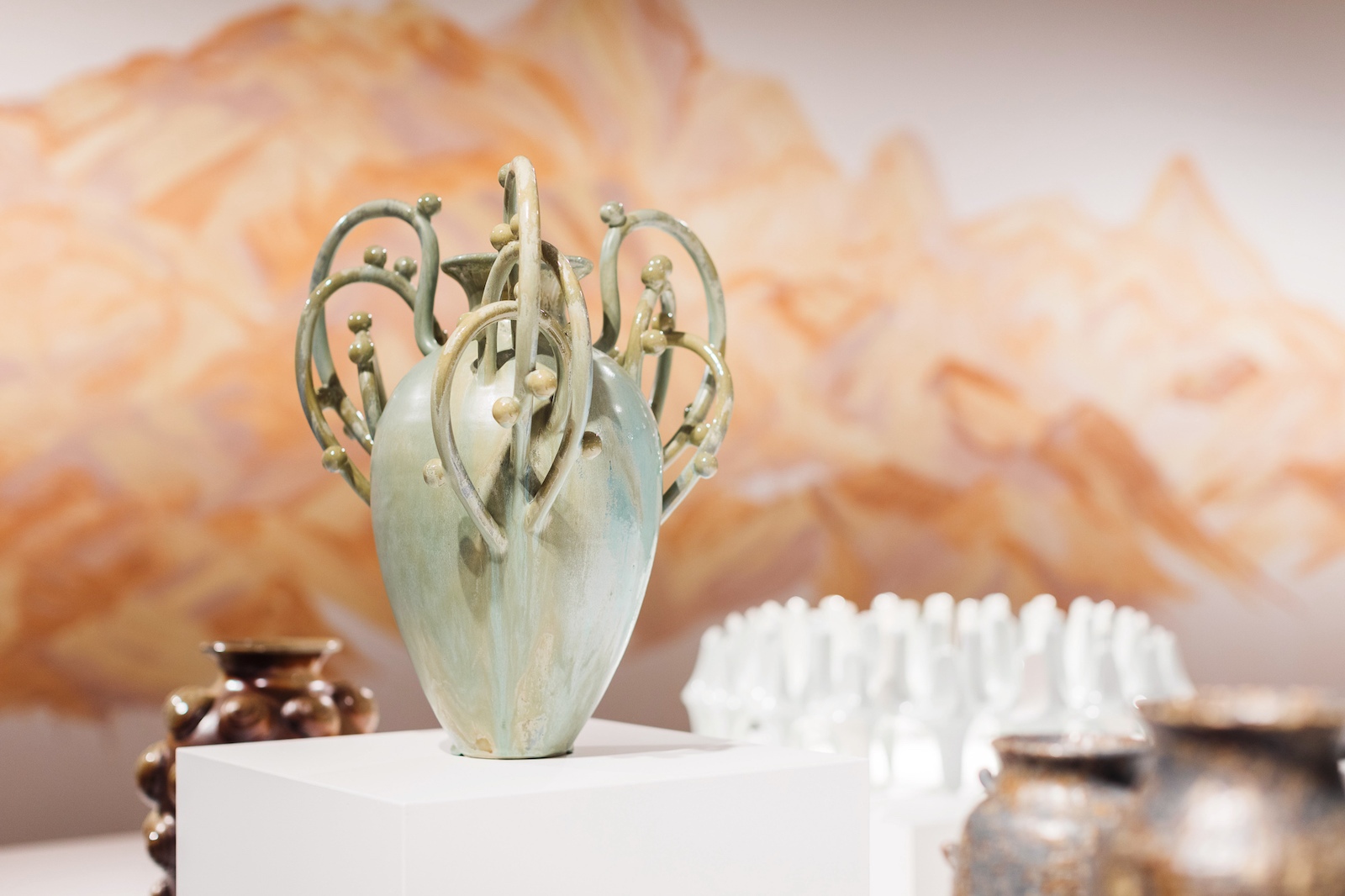 A pale green vase with multiple intricate handles sits on a plinth