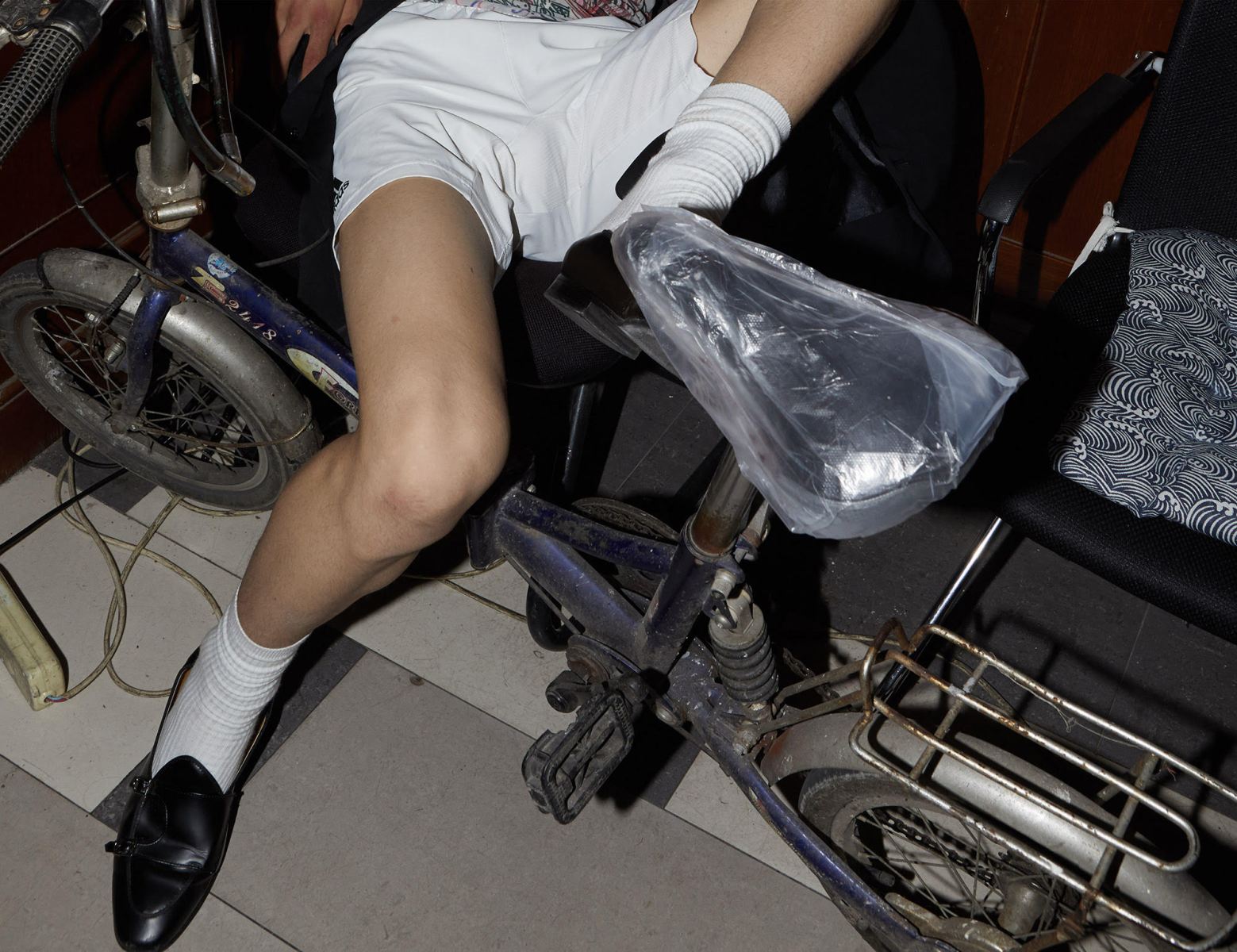 A tightly cropped photograph shows the legs of a figure sitting awkwardly over a bike, the seat is wrapped in a prominent plastic bag