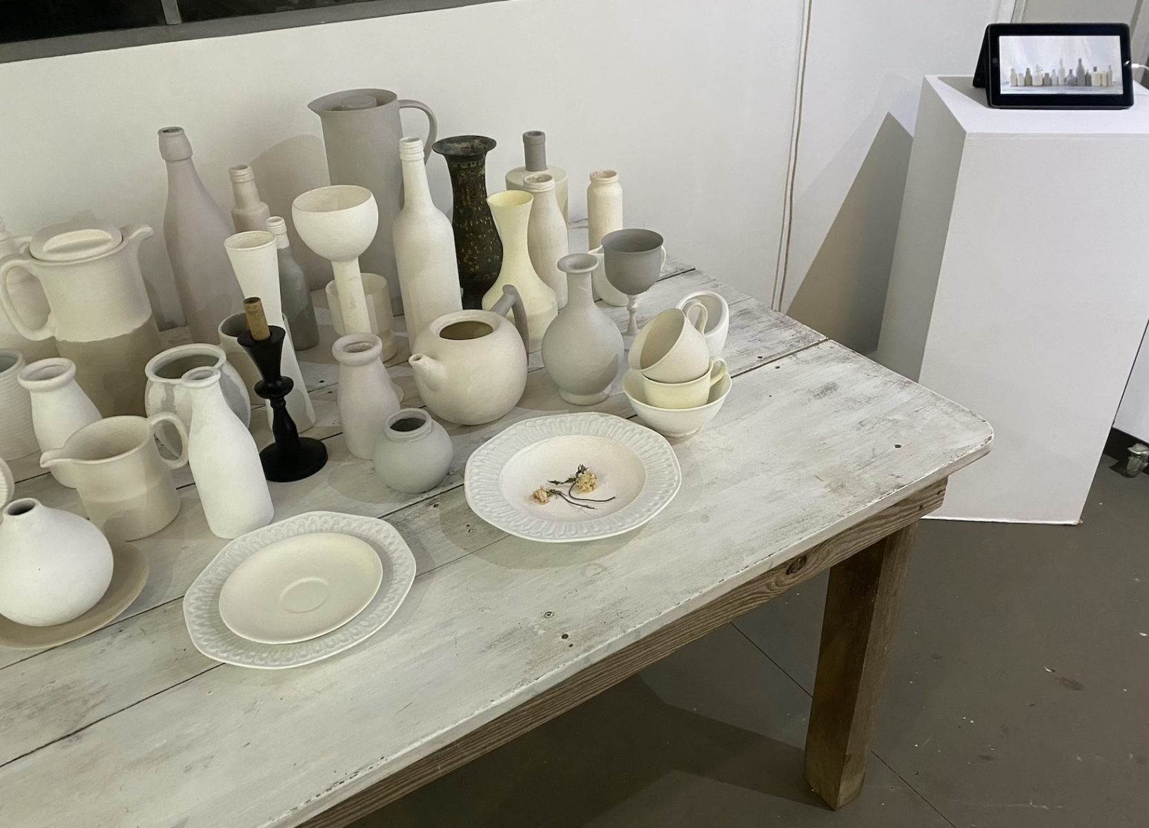 A large collection of neutral-toned pottery objects sit on a table, a video of the pottery objects runs on a tablet on a nearby plinth