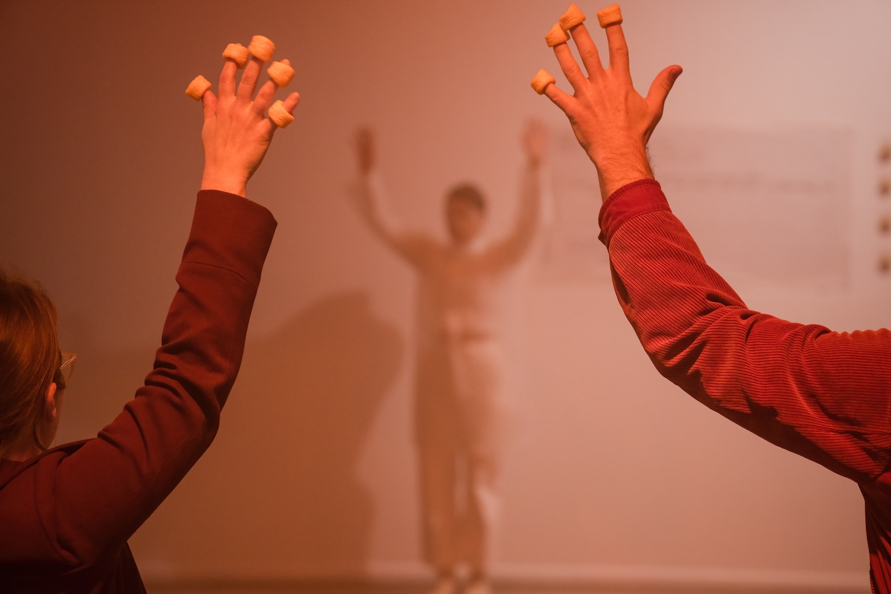 Two audience members stand side by side with their arms raised above their heads and fingers splayed. On each of their fingers is an orange Cheezel. In front of them is Merinda in white, leading them through the words and movements of 'Cheezel Rock'.