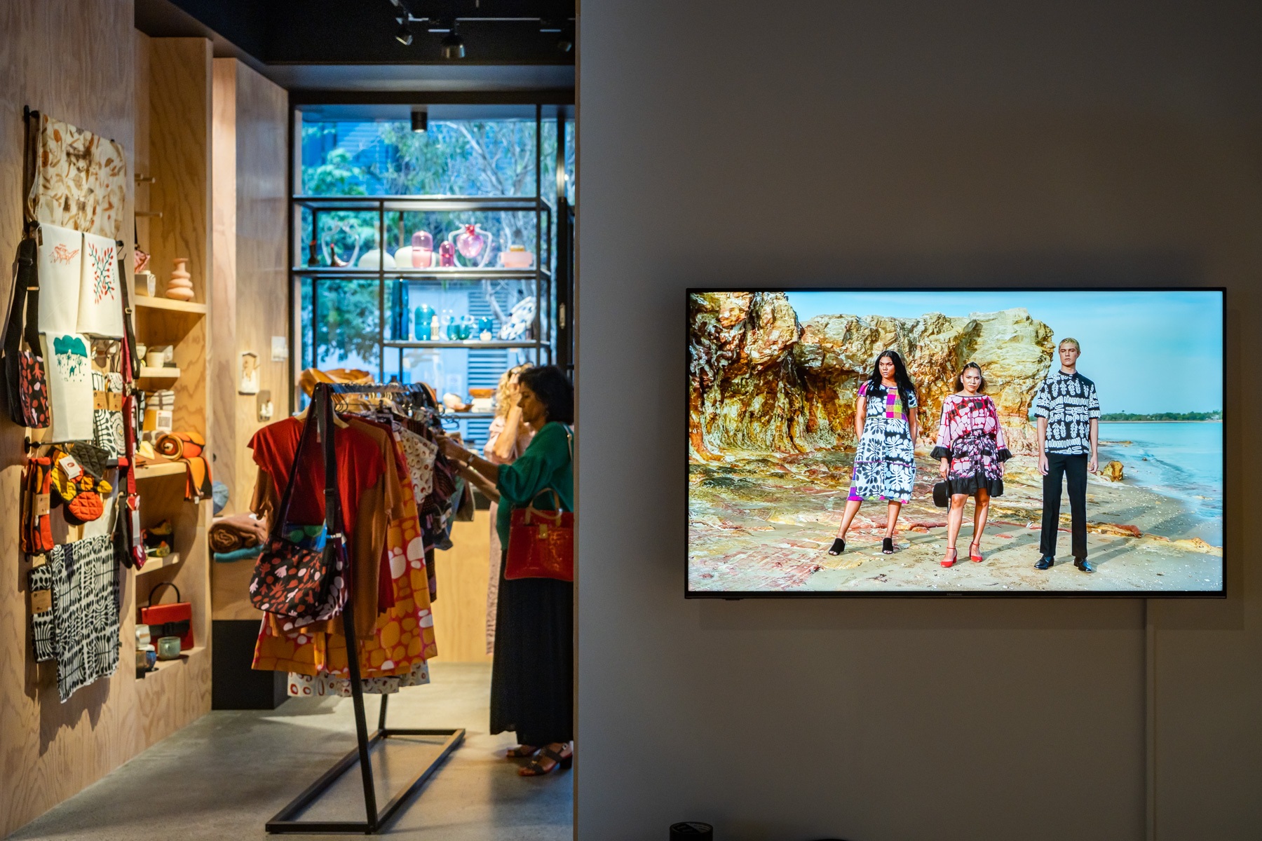 A photograph captures a woman browsing clothes in a gallery store filledl to the right a video shows three figures modelling contemporary Indigenous clothing at a striking seaside location
