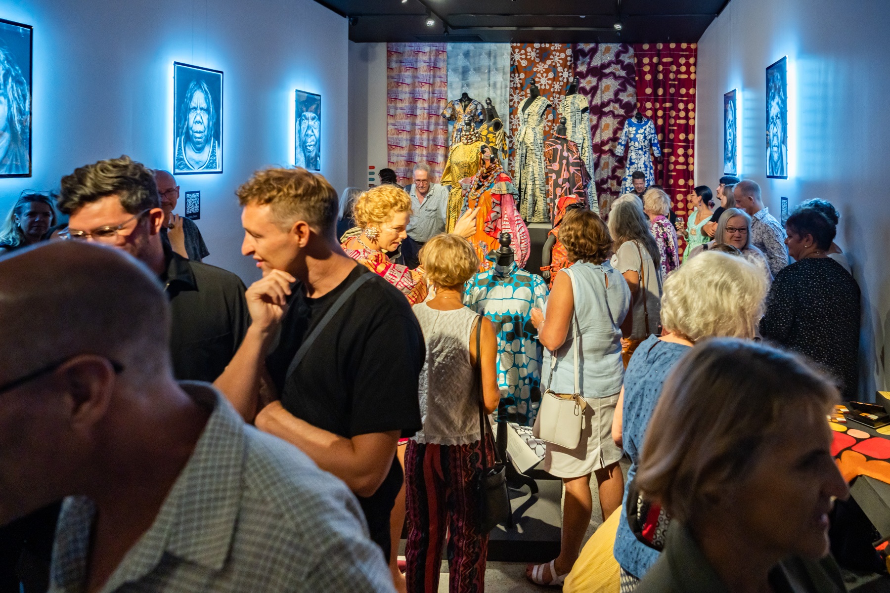A crowd fills a gallery, in the background Indigenous-designed fabrics fill a wall