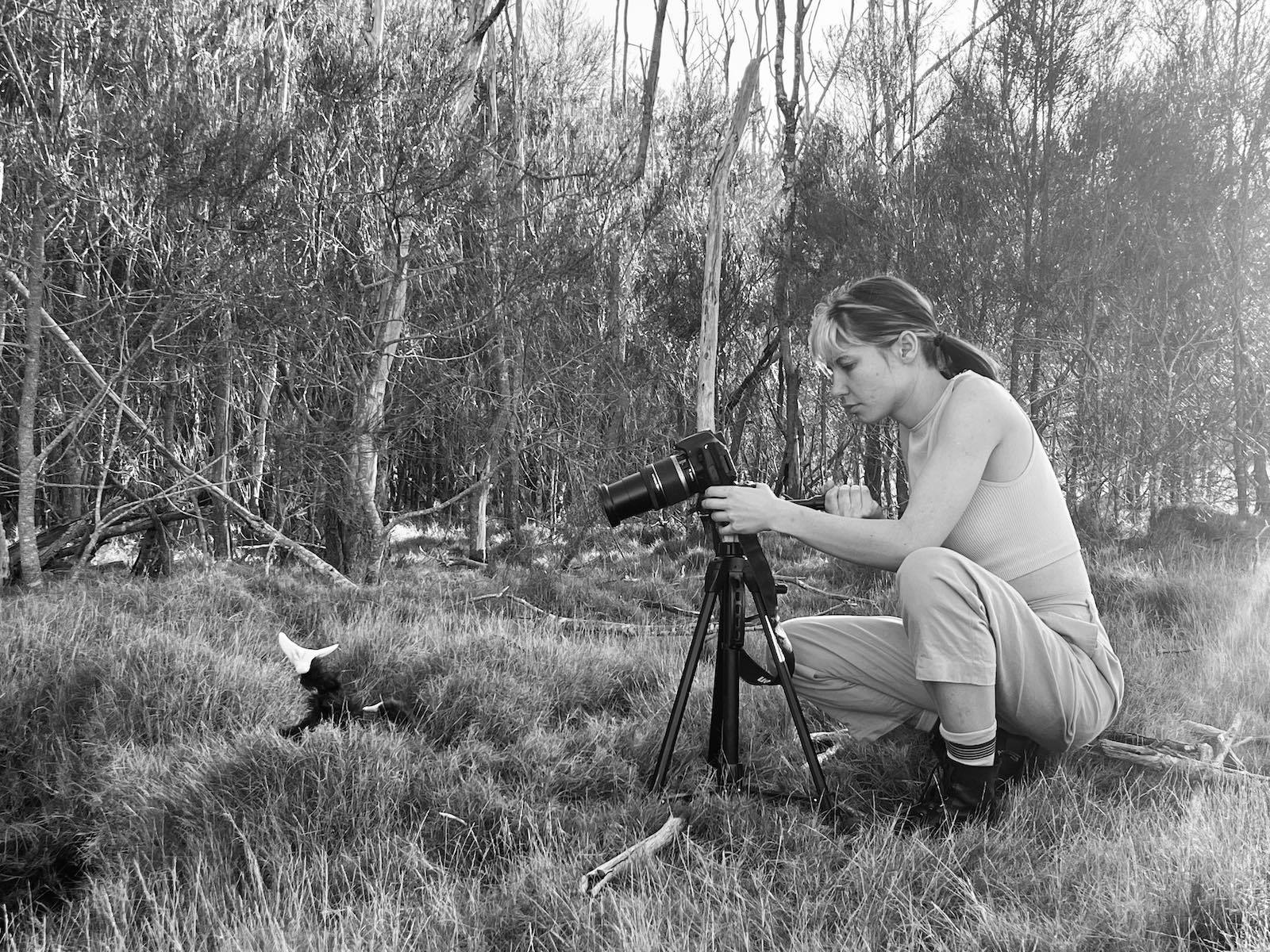 A black and white photograph captures a young person in the landscape photographing a small object
