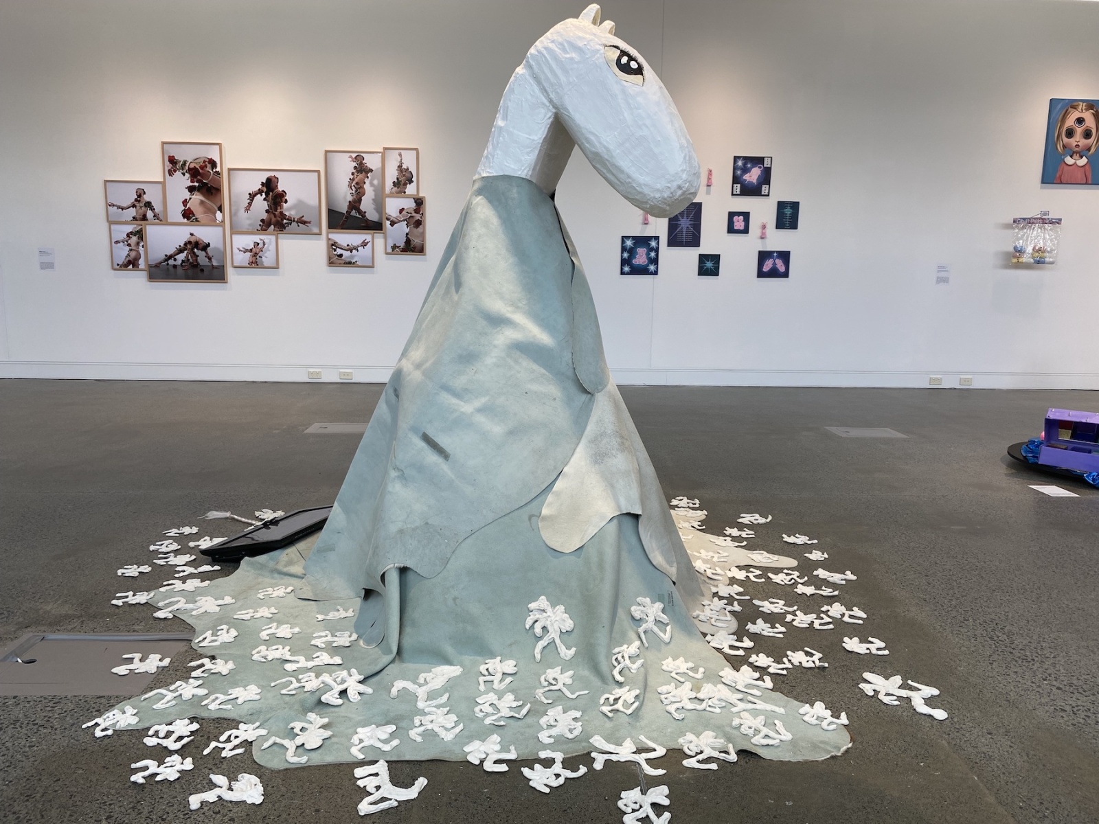 A large styrofoam horse head sits on top of a skirt made of cow hide, around the base of which sits small white plaster shapes and a television screen.
