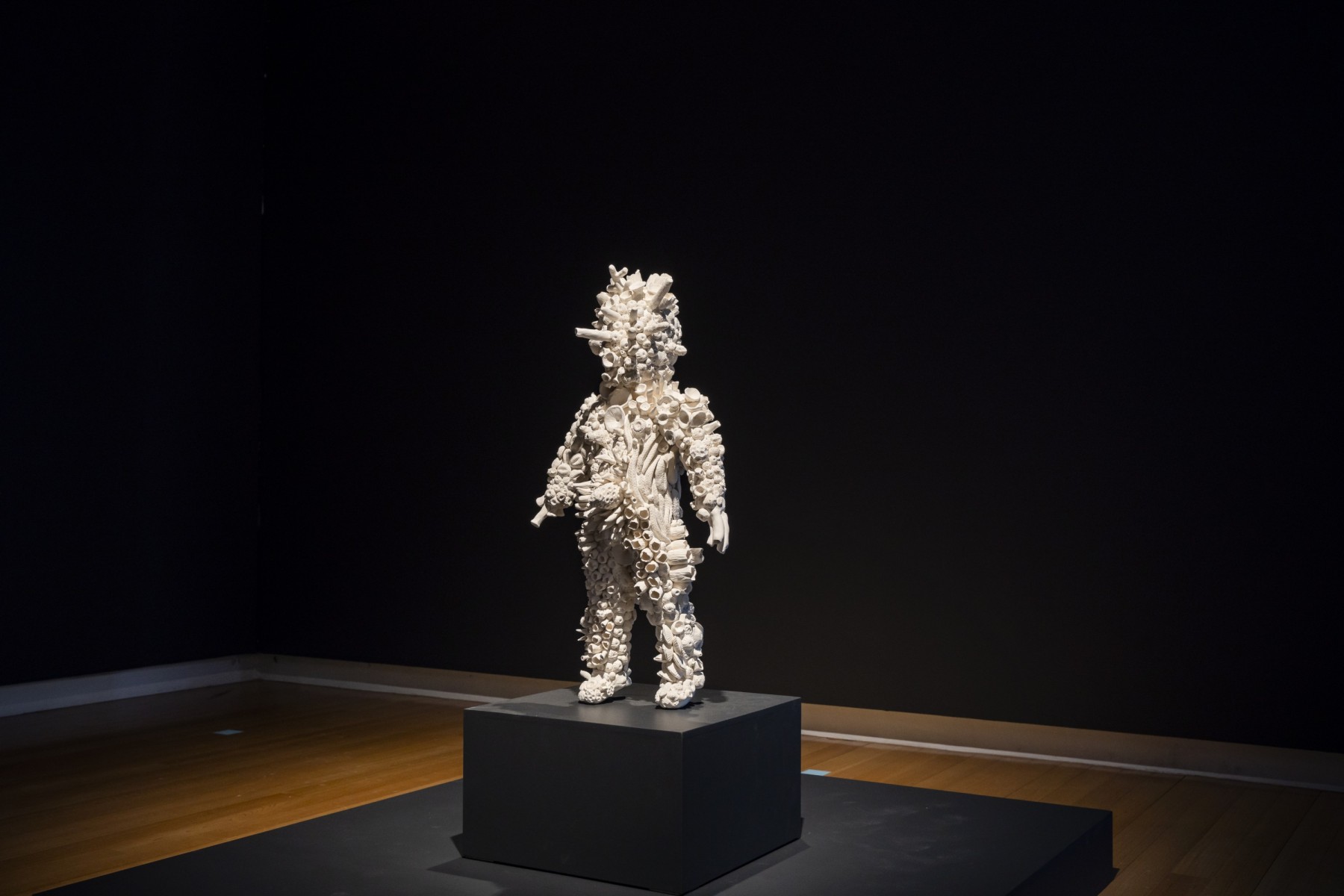 A semi-human form covered in white porcelain coral shapes stands on a black pedestal
