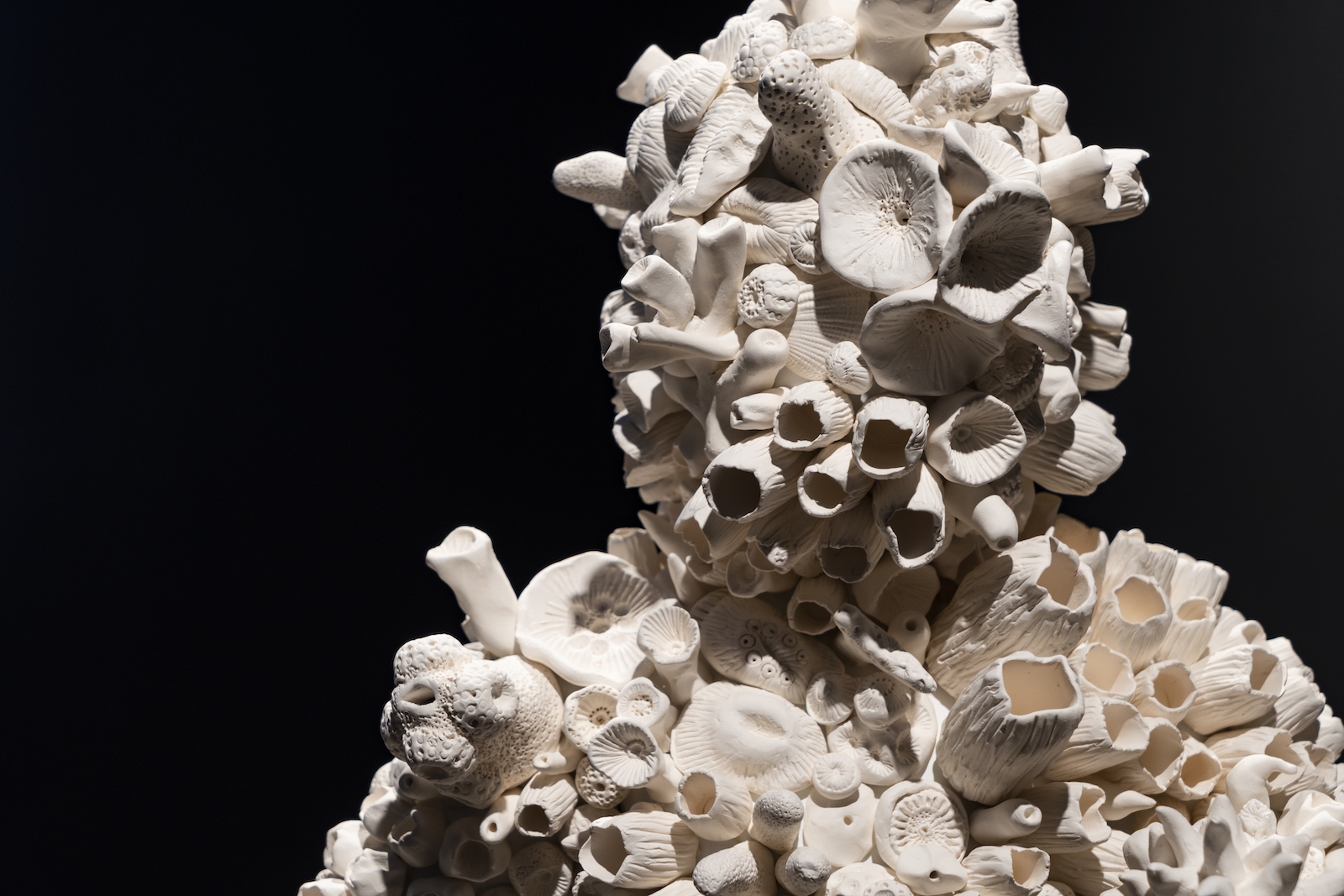 A close up shows a semi-human form covered in white porcelain coral shapes