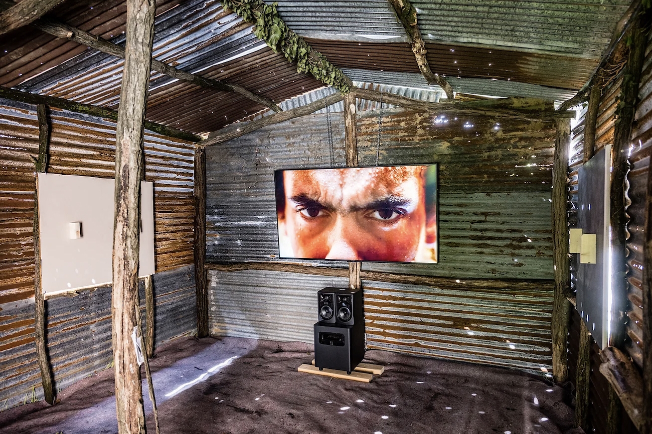 Within a tin shed, a screen shows a close up of a man’s eyes