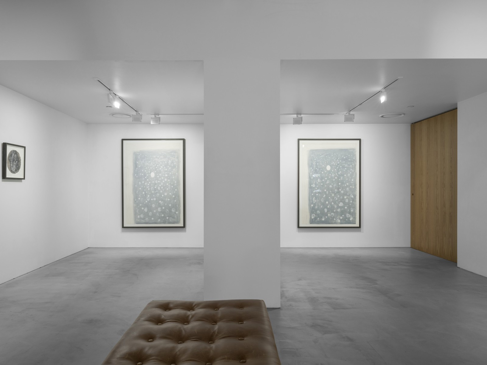 Two large and soft grey rectangular works hang upright on the far wall of a gallery, a small darker and circular work hangs to the left