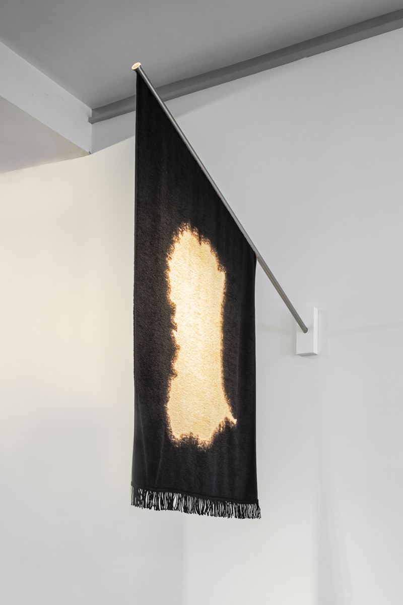 In a white gallery, a burnt black towel hangs from the wall like a flag.