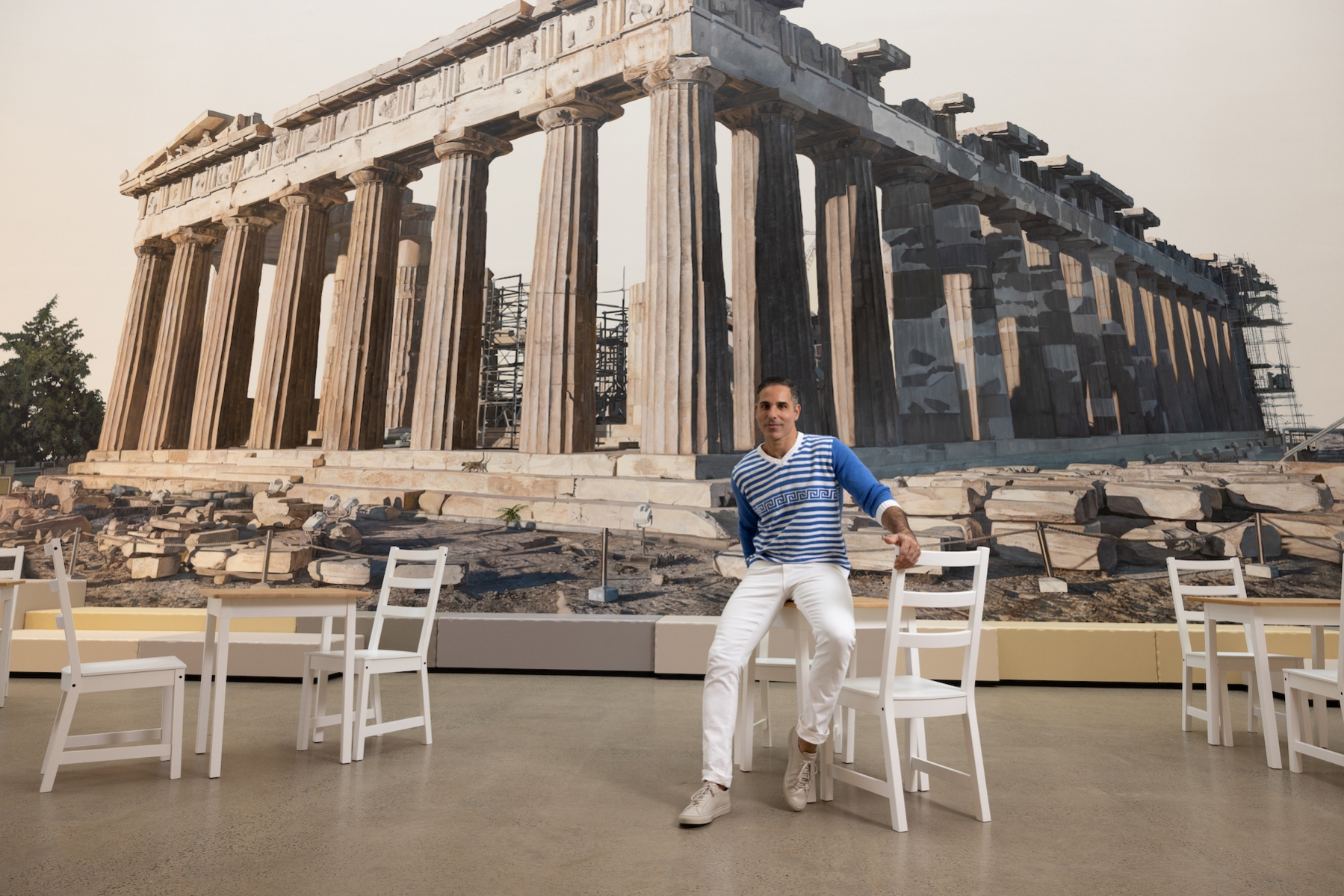 A man sits among white cafe furniture in front of a trompe l'oeil painting of the Parthenon