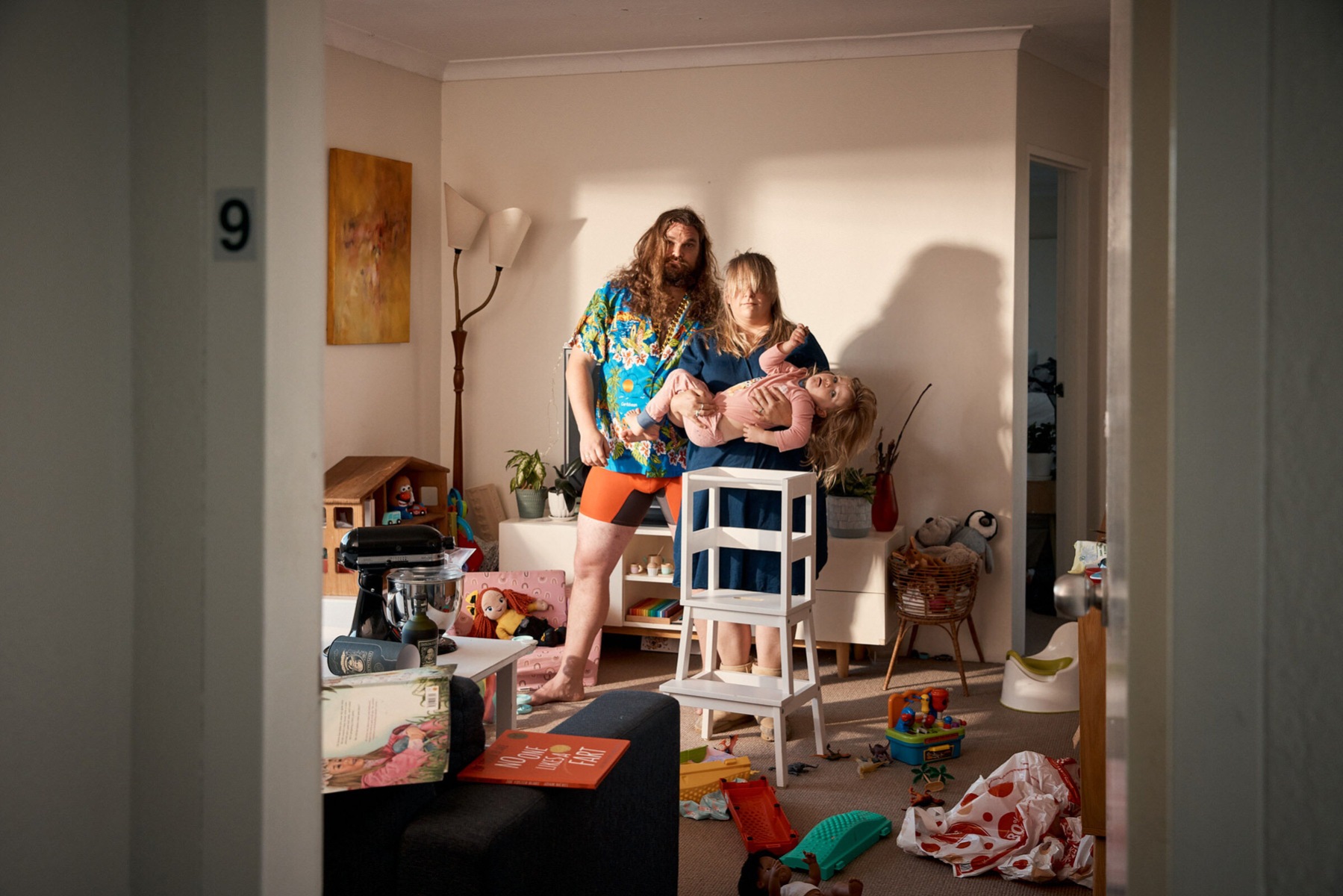A photograph of two disheveled adults and a child in a room scattered with toys, furniture, books and cookware
