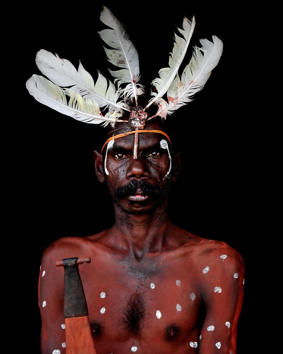 portrait of an Indigenous person wearing a feathered headdress and body paint, and holding a tool