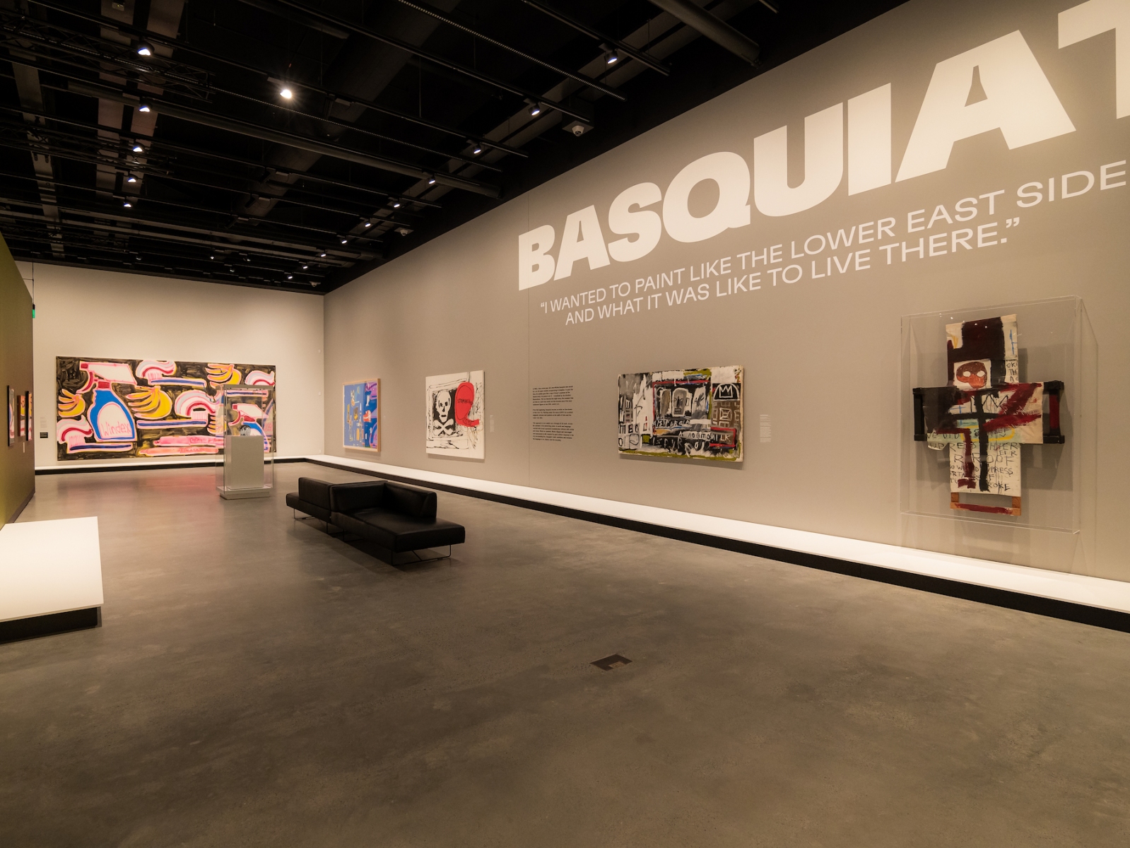 A gallery splashes the name Basquiat in large caps lock letters along a wall, underneath hang paintings by the artist