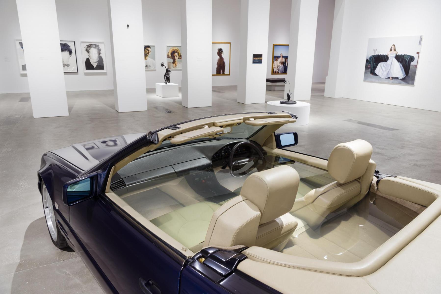 A car filled with water is parked in a gallery; long white coloumns partially block painted portraits on the far wall
