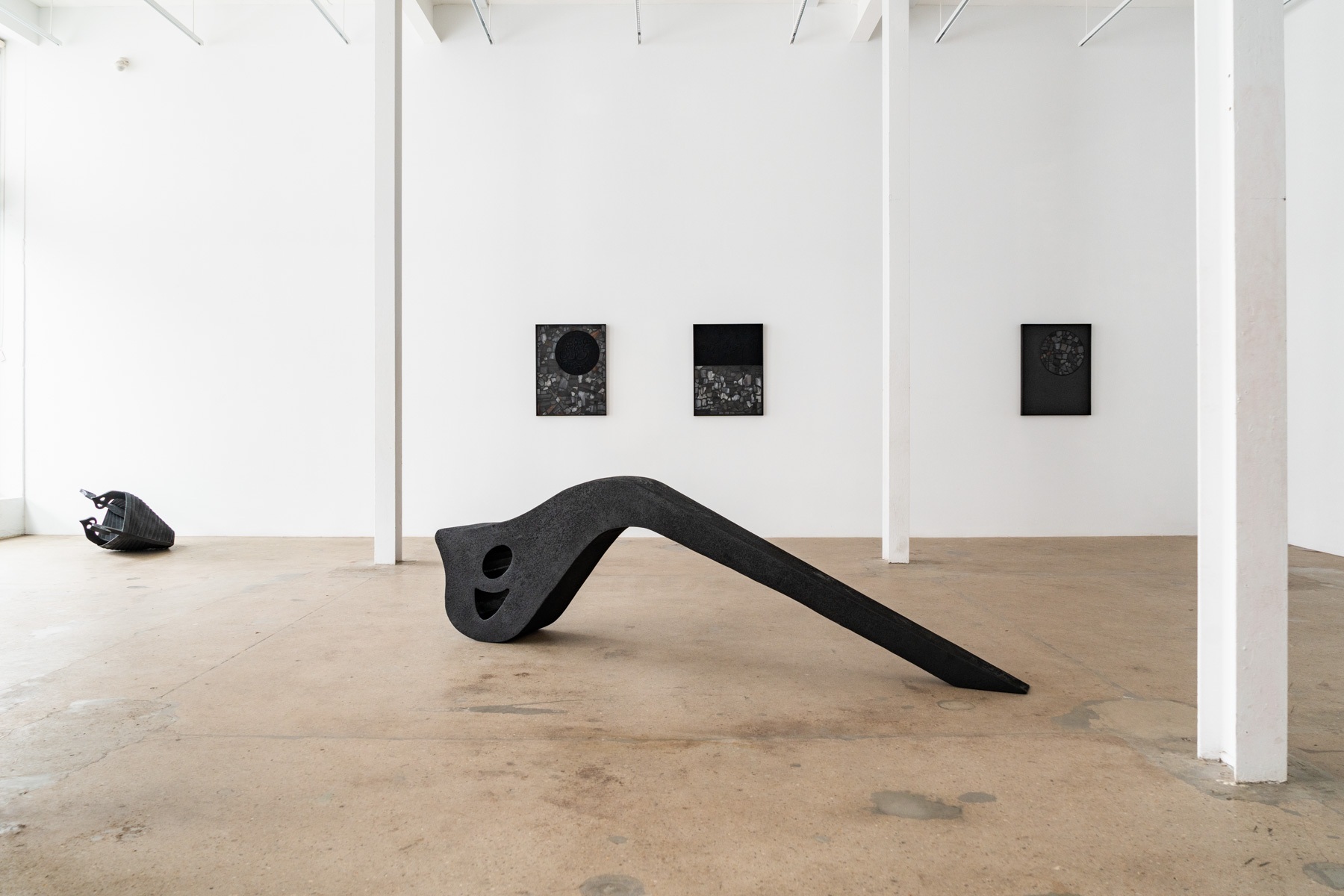 In a gallery, two large dark sculptural forms: one like a boat and the other like a lettered symbol lie on the floor. Three small dark rectagular works hang on the far wall.