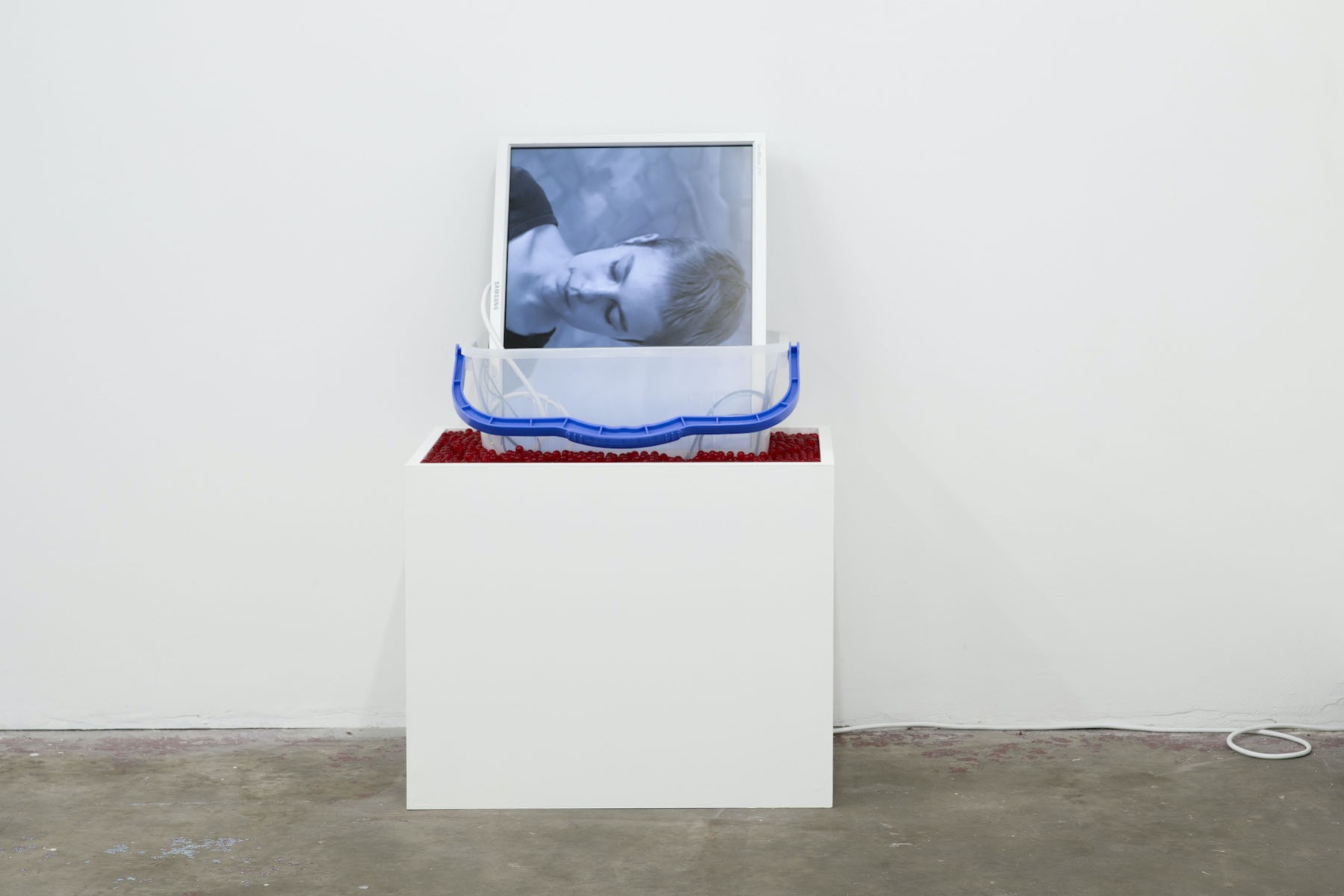 A screen showing the artist's head sits sideways in a plastic bucket. The bucket is surrounded with small red beads and sits atop a white plinth
