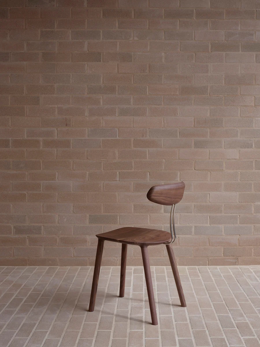A simple and minimalist ash wood chair stands against a plain brick wall and flooring.