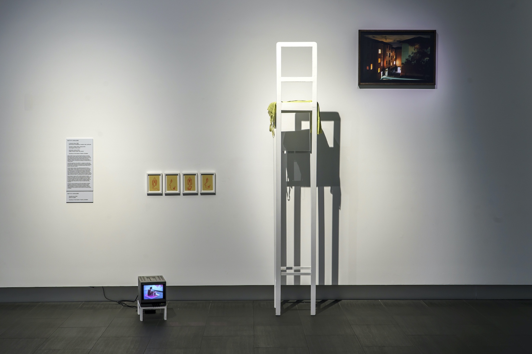 A collection of works including a very small screen on a small plinth, orange drawings of a house, an extremely high white chair, and a photograph of a building at night are arranged along a white gallery wall
