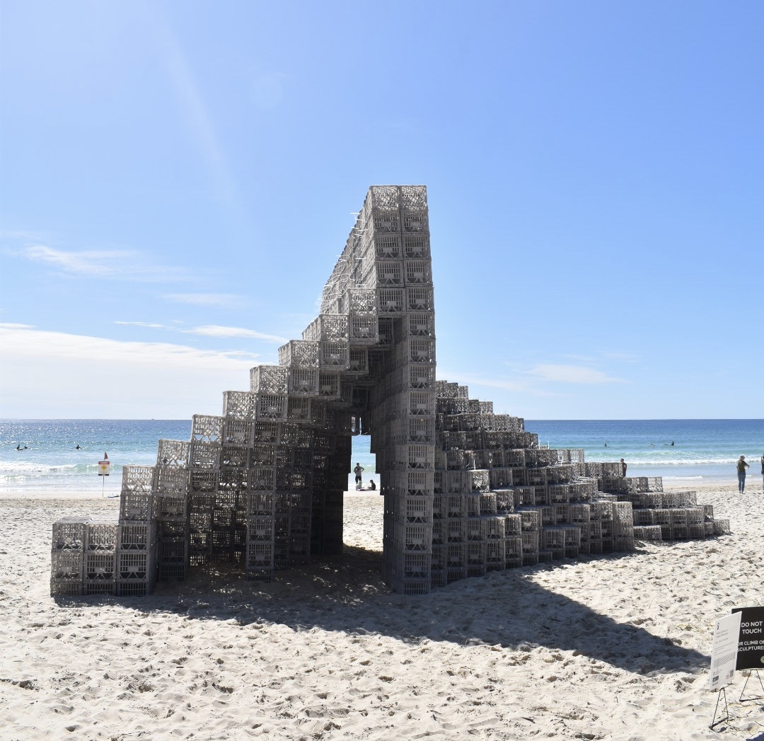 A complex sculpture made of grey milk cartons sits on the beach; in the distance is the ocean.