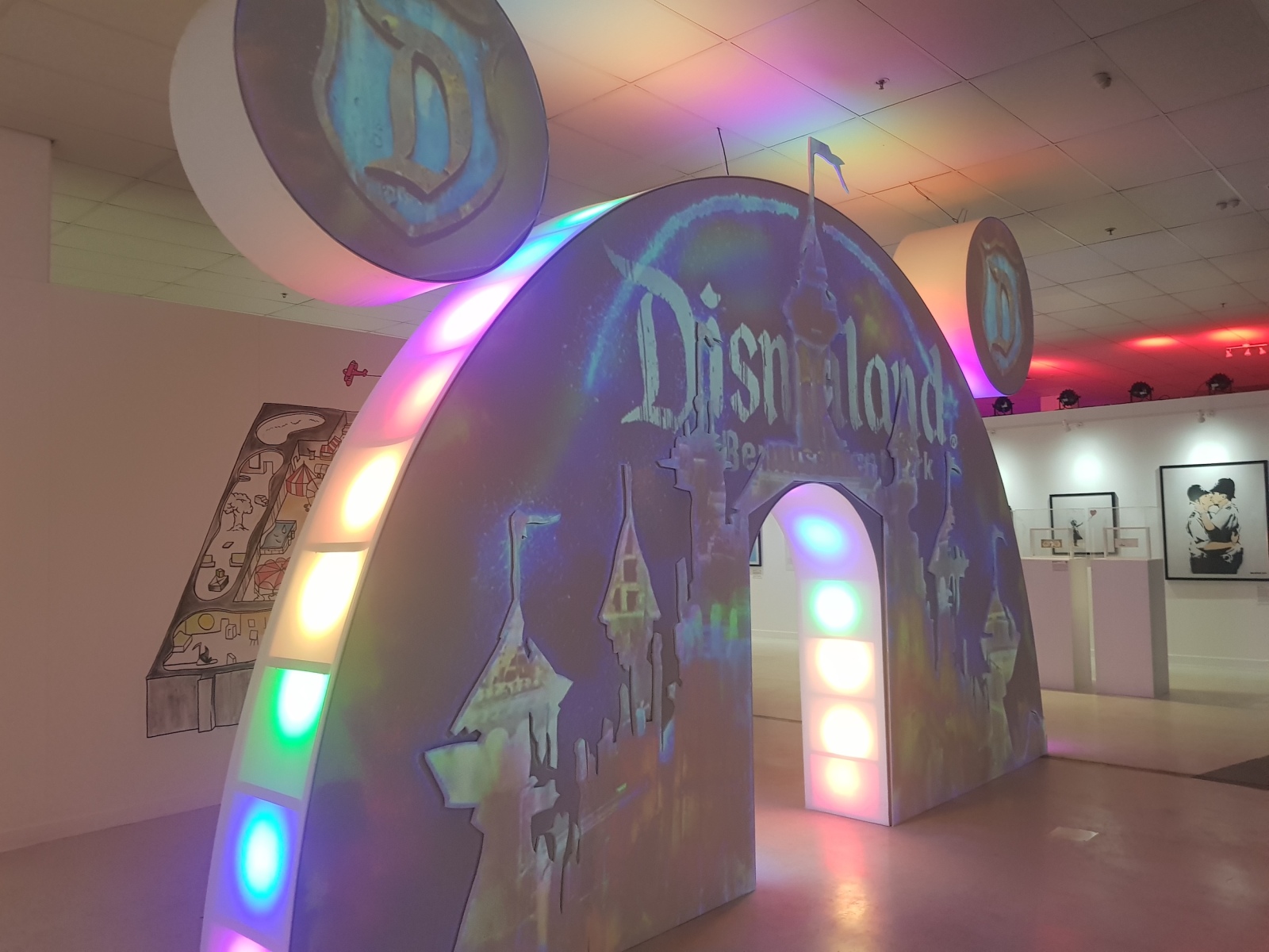 Front facing photo of a large archway reminiscent of Mickey Mouse’s ears in shape, has a projection of an edited Disney castle logo. Its sides are lit with multi-coloured LED lights.