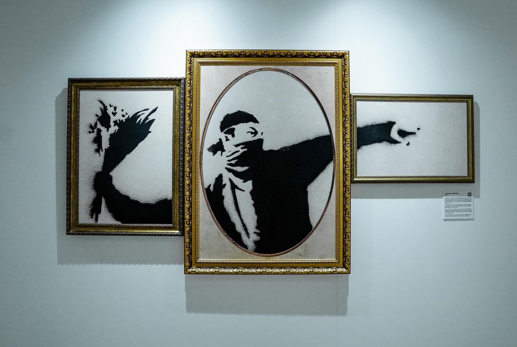 Three black and white stencil graffiti images in golden frames depict a masked man throwing flowers