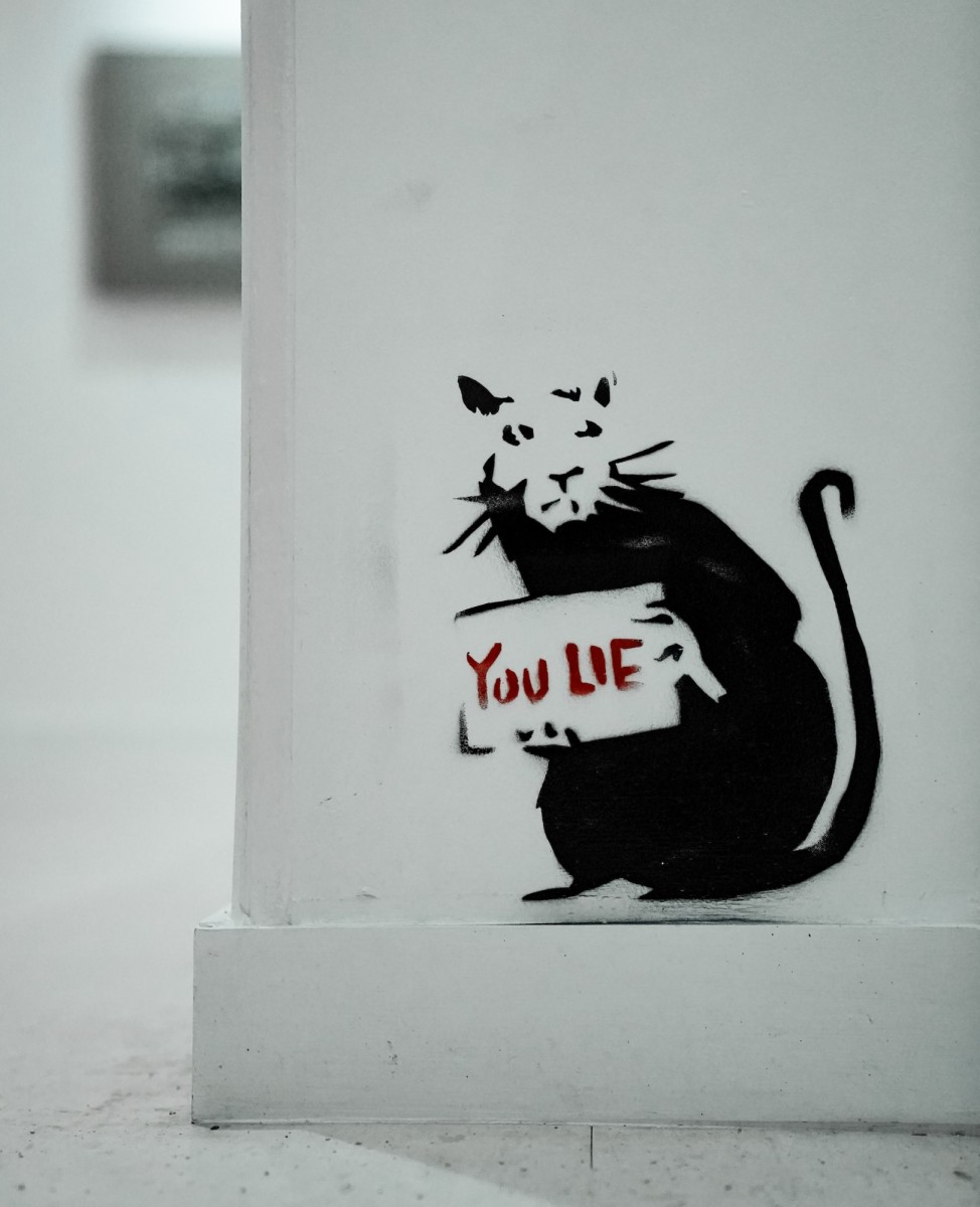 A stencil graffiti depicts a rat holding a sign with the words "you lie"