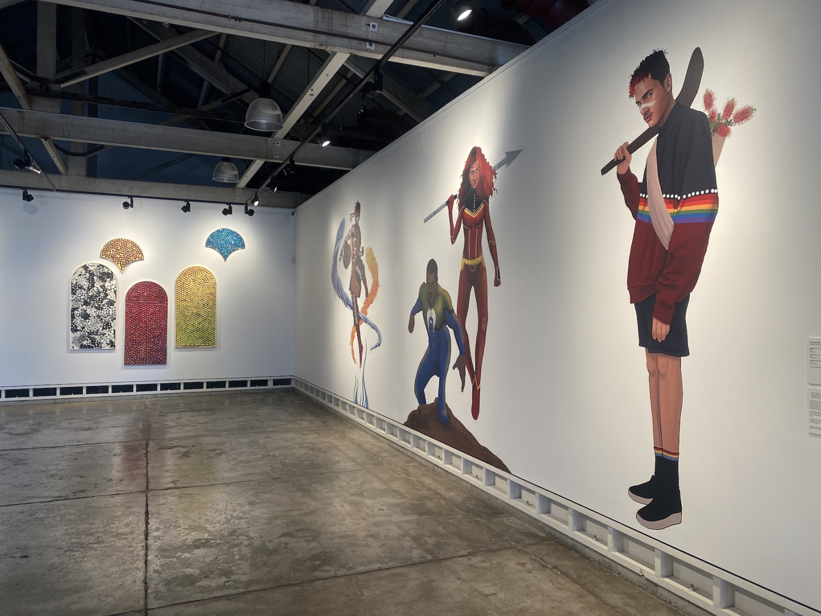On two white gallery walls hang a series of bright shapes (left) and four larger than life digital drawings of Indigenous superheroes (right)
