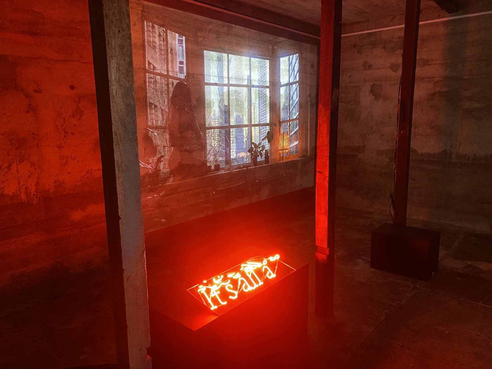 Red light from neon text reading "it's all a" fills a room of exposed timber and concrete, on the far wall a video projection shows a figure sitting in front of a city window