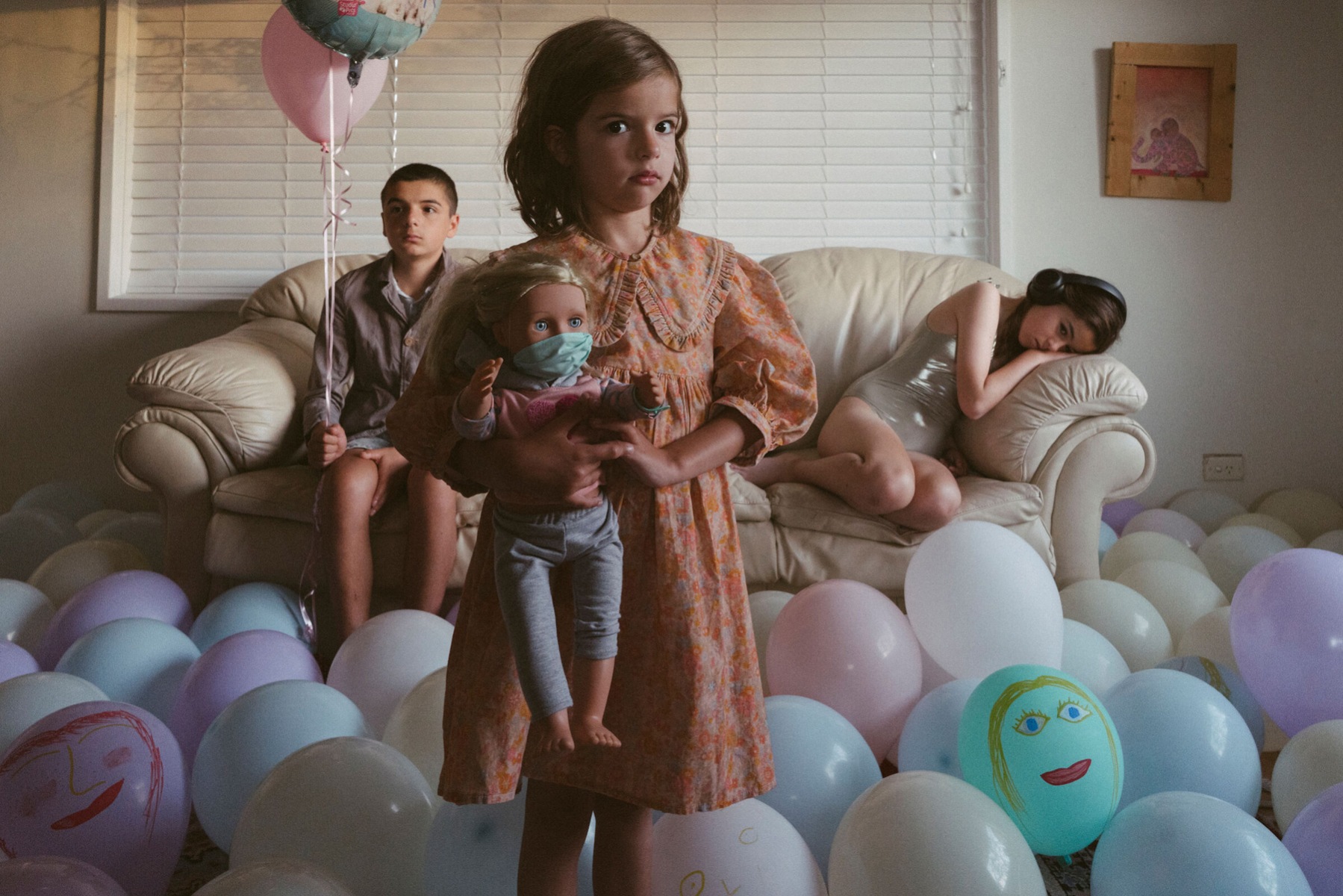 A photograph of three children in a living space surrounded by balloons, two are sitting on a couch and one is standing in the foreground, the child on the left is holding a balloon, the child on the right is wearing headphones and the child in the centre is holding a doll with a makeshift facemask