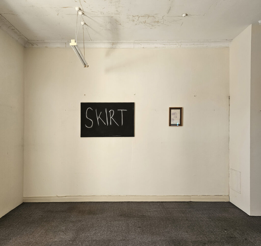Two artworks hang in an old and otherwise empty room, one has large capitalised letters reading ‘skirt’ on a blackboard. 