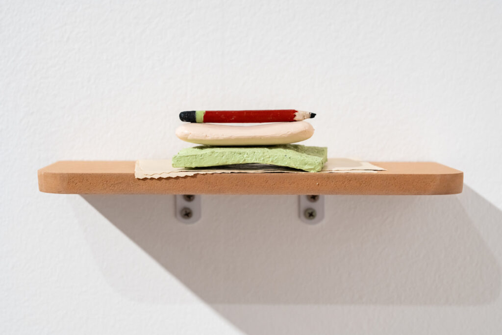 A timber shelf with a piece of cloth, green sponge, bar of soap and pencil stacked on top of each other. 