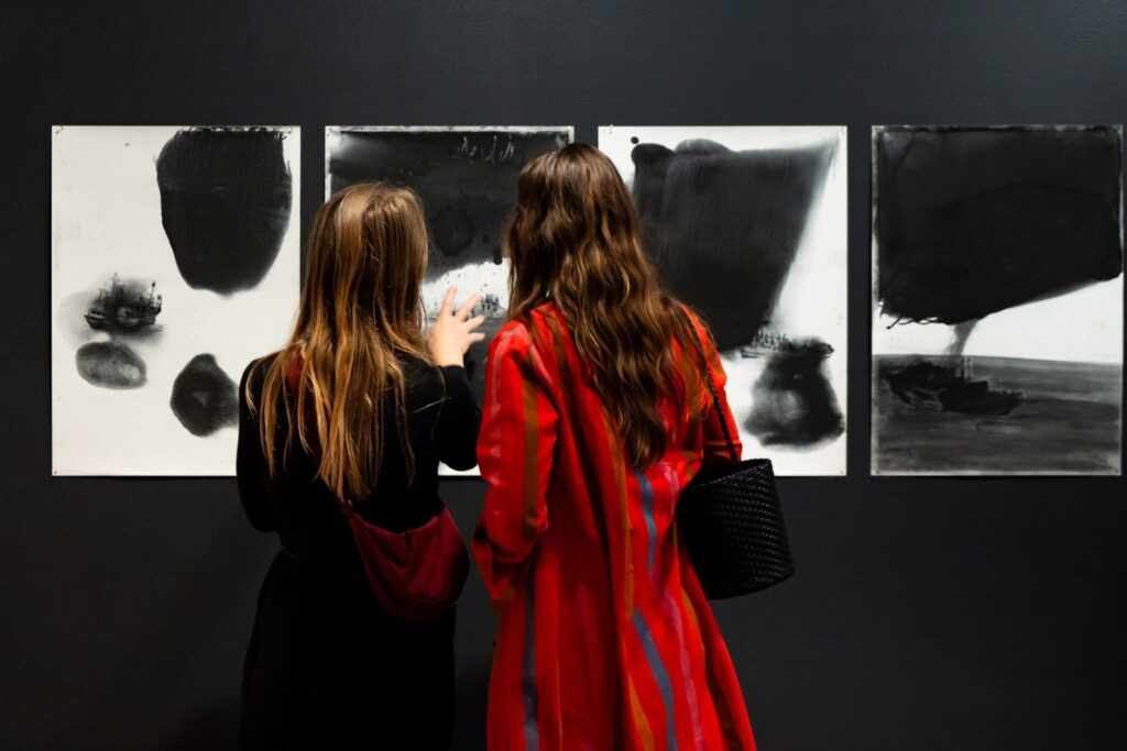 Two women stand in front of four works on paper. Each features soft rounded black forms against white.