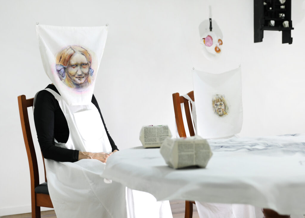 A woman wearing a white apron sits at a dining table. Over her head is a pillowcase painted with the face of a young girl. We can only see the figures hands.