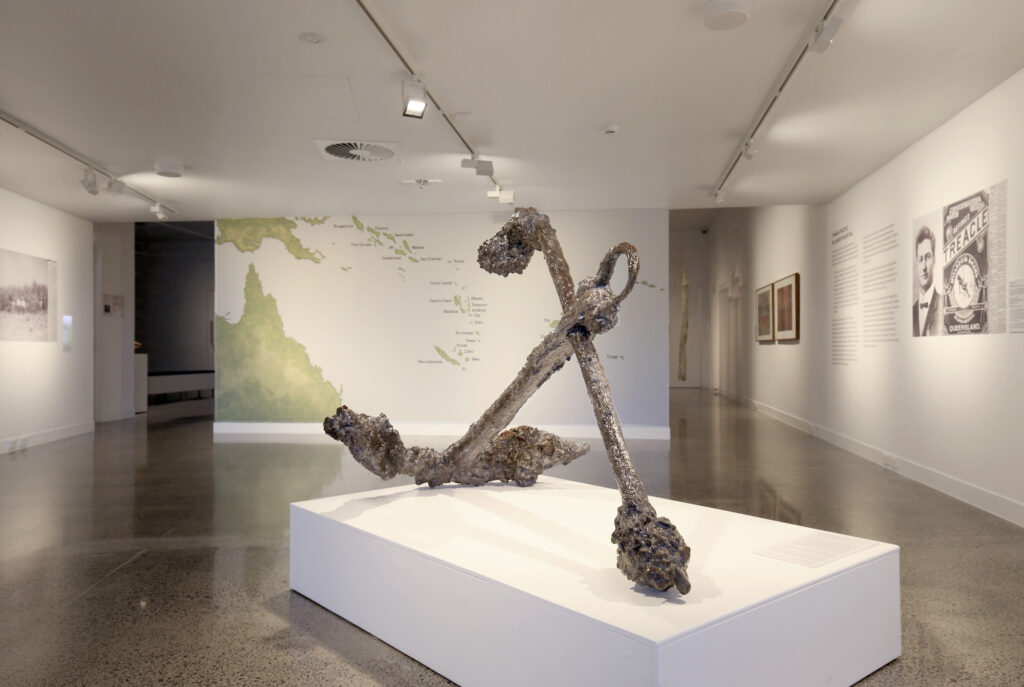 A gallery with black-and-white photographs on the walls, a large colourful map of Australia and the South Pacific, and a large rusted anchor in the foreground.