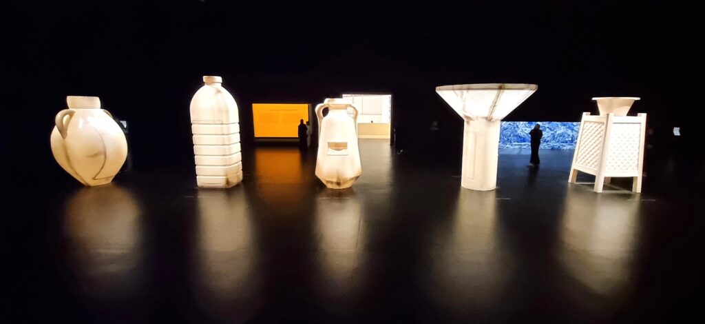 Plastic vessels made larger than human are lit from within in a dark gallery space