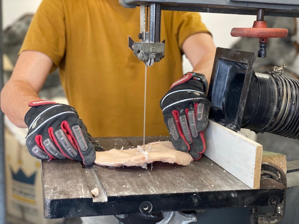 A close up of a person in a mustard coloured T-shirt, wearing black protective gloves with red and white trim. Their face is not visible. They are holding a small waxy, flesh coloured object on a bench as it is cut in half by a bandsaw.