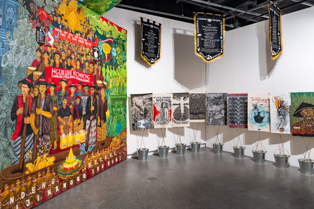 To the left, a bright and dense mural hangs in a gallery; to the right are small placards held in silver buckets; three black, white and gold banners hang overhead.