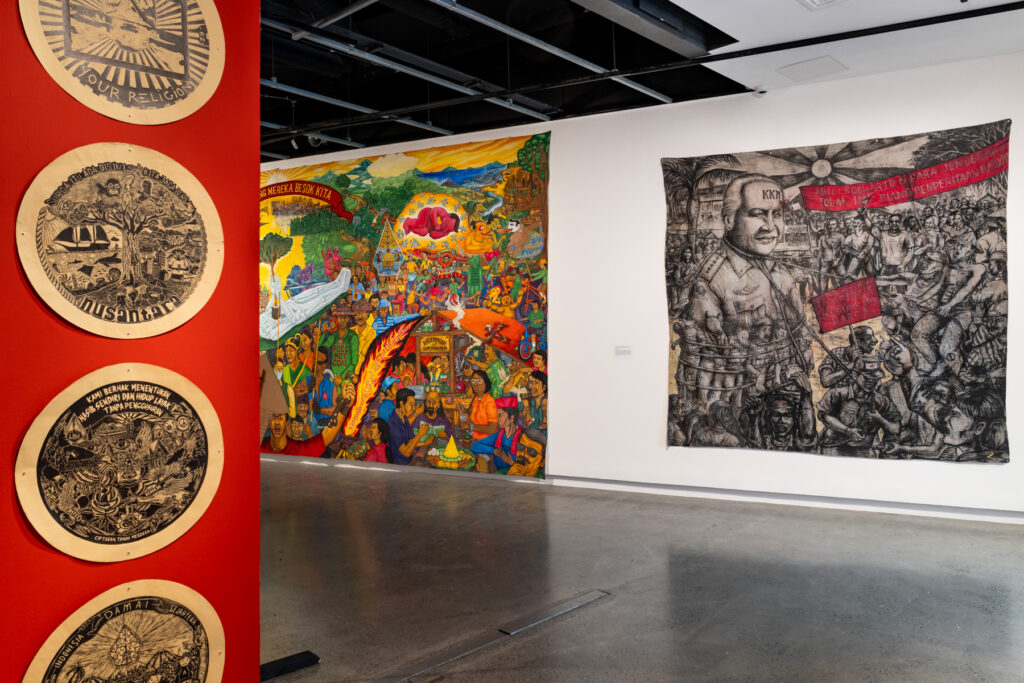Two dense and colourful mural-banners hang on a gallery wall, in the foreground round prints hang on a red wall.