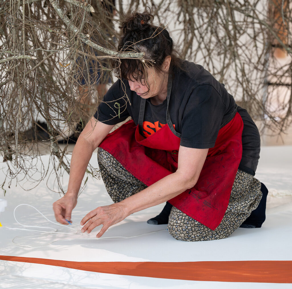 A woman installs an artwork. Behind her hang sticks, beneath her is a white ground with a thin orange-red sweep of colour.
