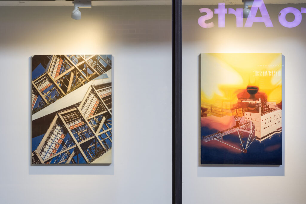 Two paintings hang in the front window of a gallery. The painting on the left features zigzagging staircases; the painting on the right features an upside down industrial scene distorted with washes of yellow, orange and pink.