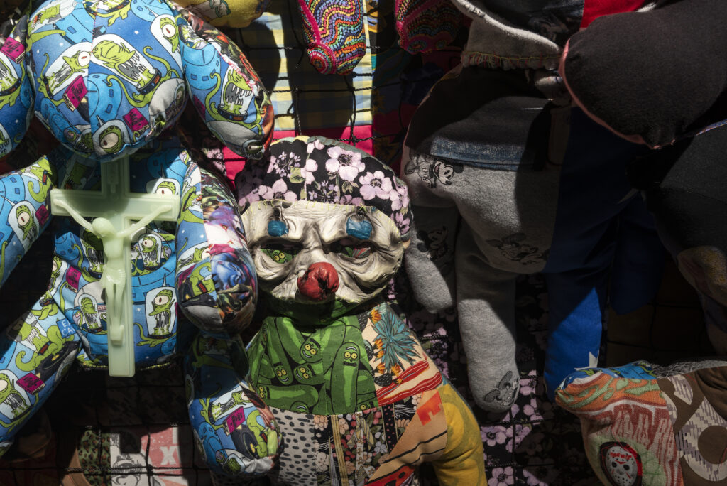 A close up image of grouped soft toy-like sculptures on a wall, one bearing a green crucifix on top of an alien cartoon printed fabric toy next to a clown like soft sculpture.