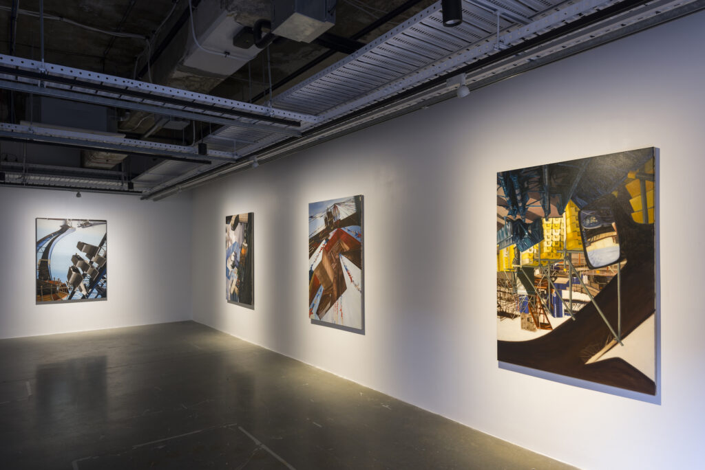An installation image of a gallery, with four industrial paintings on display. The walls are white and the ceiling is exposed. 