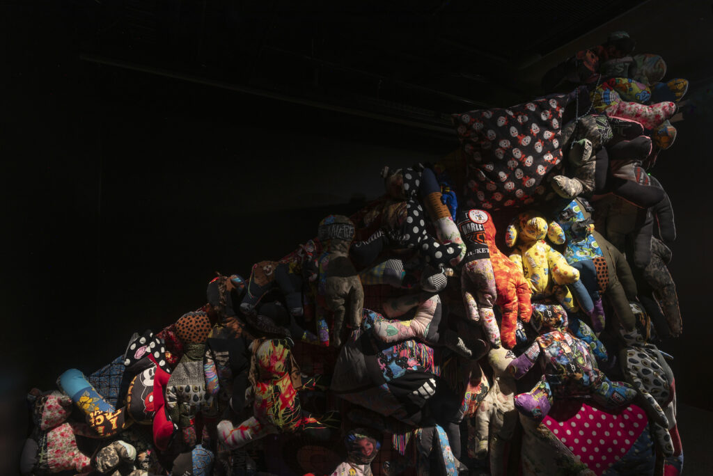 An image of a dark room displaying the side view of the installation, dimly lit with colourful soft toy-like sculptures tapering diagonally from top to bottom.