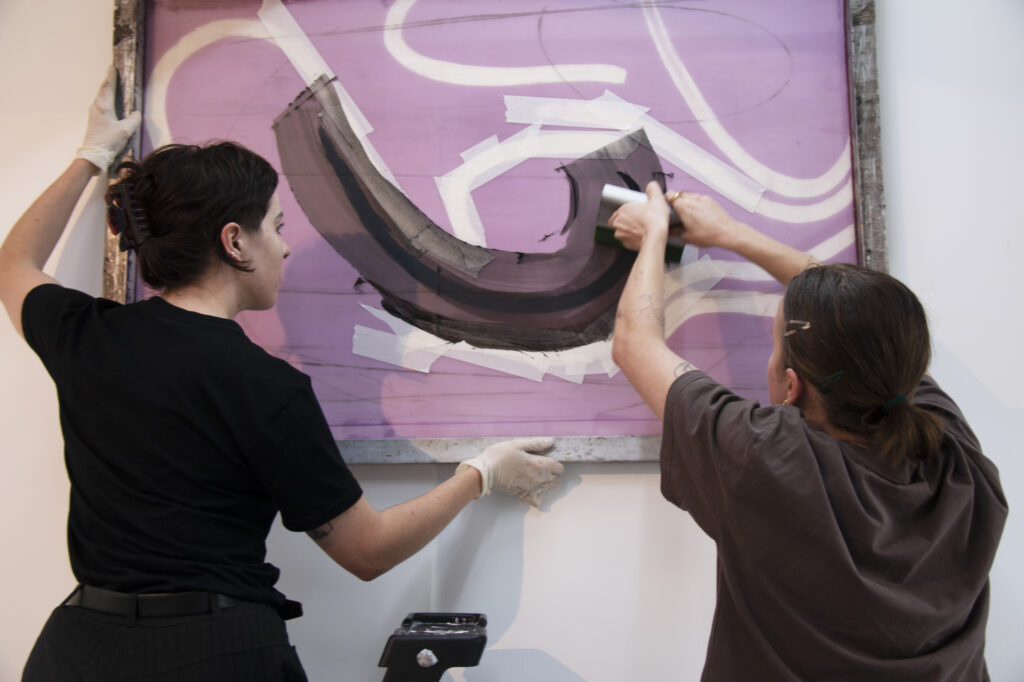 Two figures, Isabelle Cowan and Stella Haycock, stand in black clothing holding a large silkscreen against a wall. The figure on the right uses a squeegee to draw black ink across the screen. 